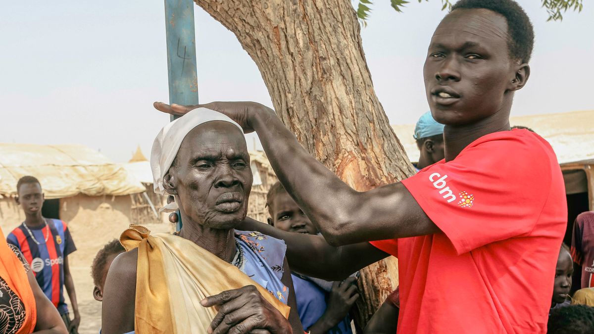 Ein junger Mann im rotem T-Shirt mit CBM-Logo misst mit einer Holzlatte die Körpergröße einer älteren Frau.