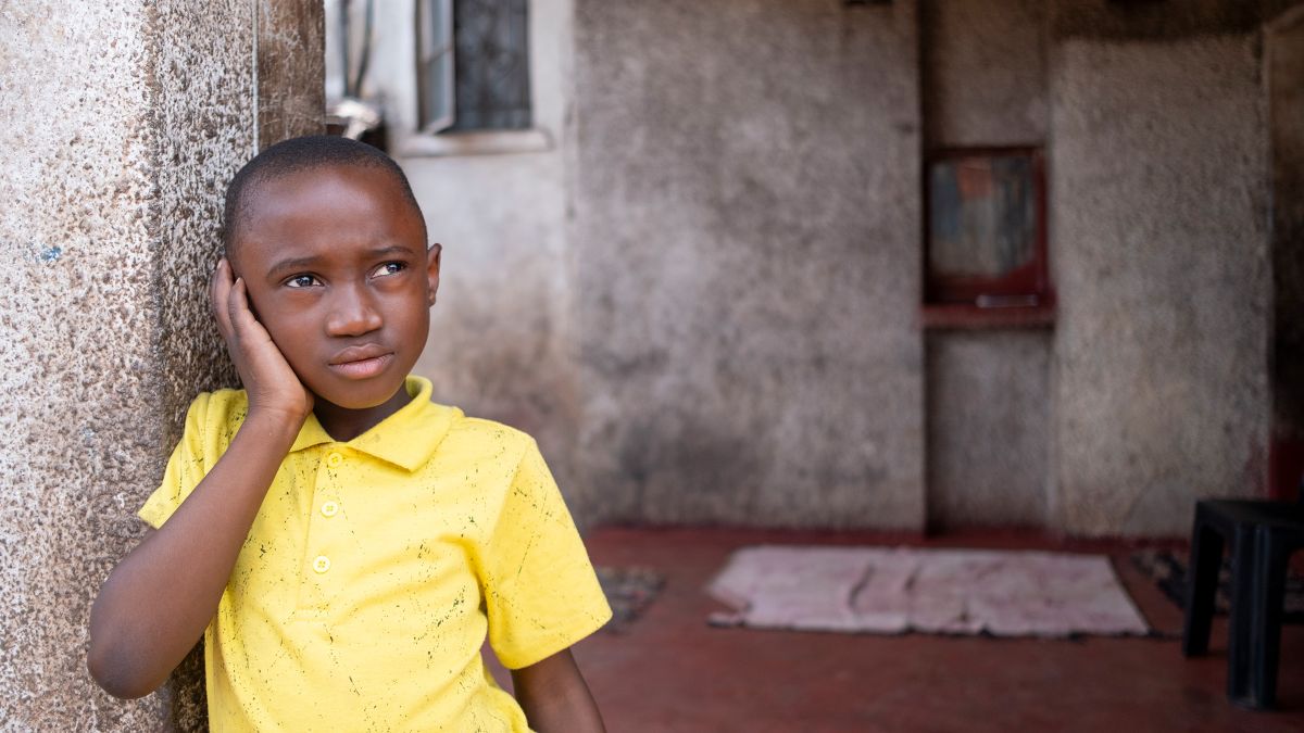 Ein etwa achtjähriger Junge lehnt sich an eine Verandamauer; erhält seine rechte Hand an sein rechtes Ohr.