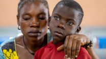 Eine Frau mit halb geschlossenen Augenlidern legt ihren Arm um die Schultern eines Jungen mit weiß scheinenden Pupillen.