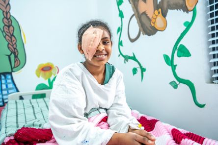 Ein etwa elfjähriges Mädchen mit Augenverband sitzt auf einem Bett. Es lächelt.