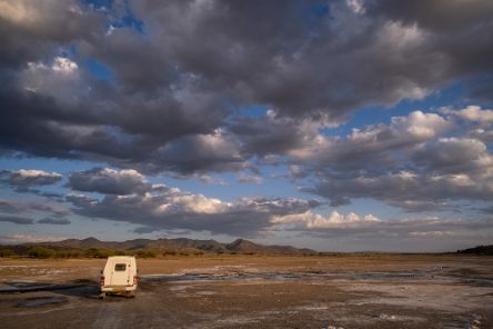 Ein Auto in savannenartigem Gelände, darüber ein weiter Wolkenhimmel.
