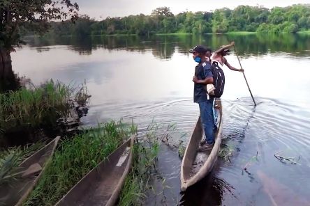 Zwei Männer stehen auf einem schmalen Boot auf einem Fluss, der von Buschwald gesäumt ist.