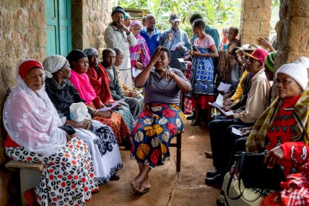 Menschen sitzen und stehen in einem überdachten Wartebereich. In ihrer Mitte sitzt eine Frau auf einem Stuhl und hält sich ein Auge zu.