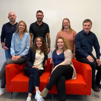 Zwei Frauen sitzen auf einem roten Sofa, dahinter stehen zwei weitere Frauen und drei Männer