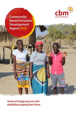 Zwei Frauen und ein junger Mann mit Downsyndrom stehen vor einer Lehmhütte auf Stelzen. Sie lächeln.