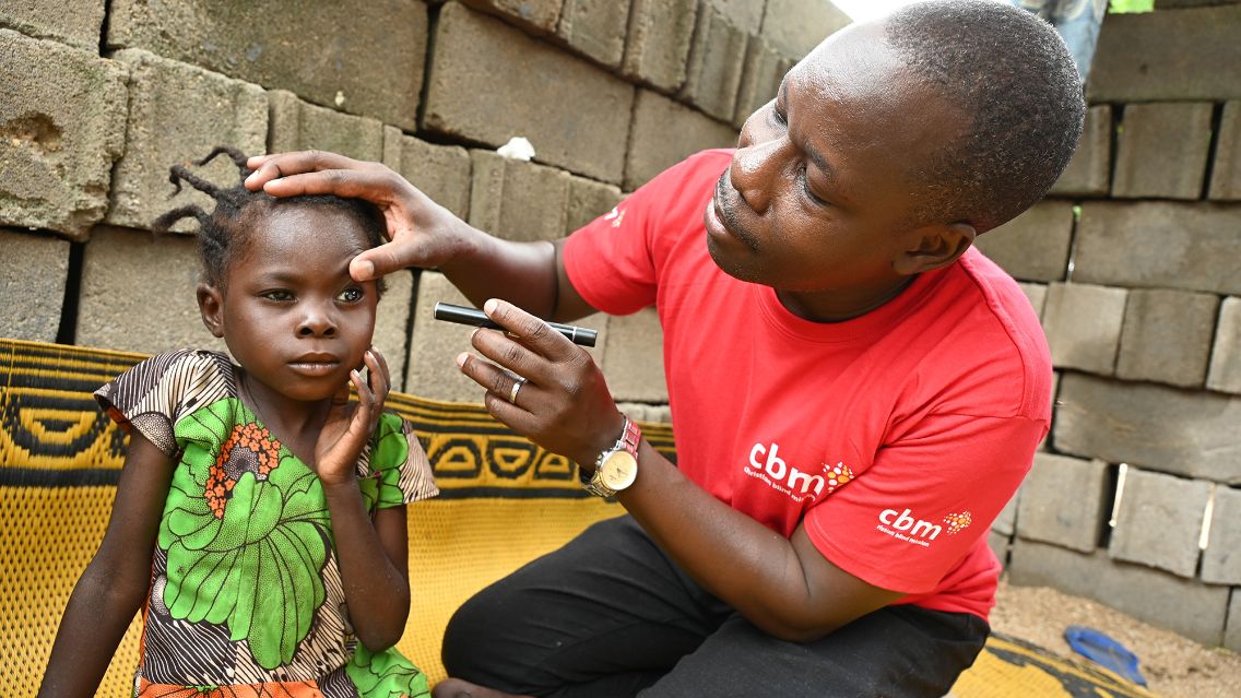 Ein Mann im roten CBM-Shirt untersucht mit einer Handlampe die Augen eines kleinen Mädchens.