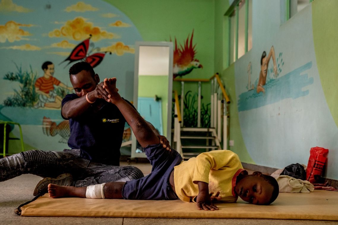 Ein Junge liegt auf einer Matte und streckt ein Bein hoch. Ein Mann hilft ihm dabei, stützt das Bein mit den Händen.