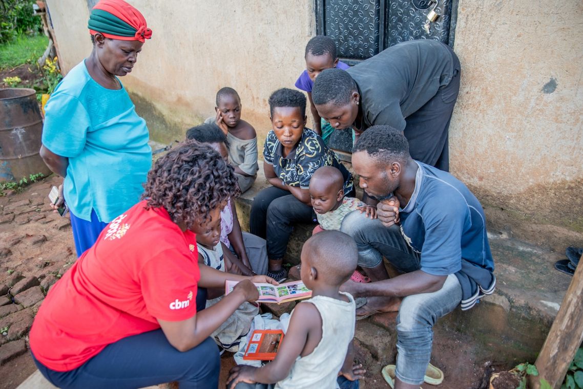 Eine Frau sitzt vor mit Erwachsenen und Kindern vor einer Hütte und zeigt einem Kind eine Abbildung.