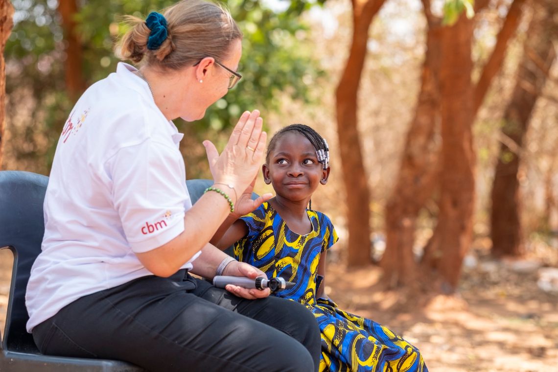 Ein etwa Neunjährige und eine Frau mit weißem CBM-Polohemd sitzen nebeneinander auf Plastikstühlen und klatschen je die rechte Hand aufeinander.