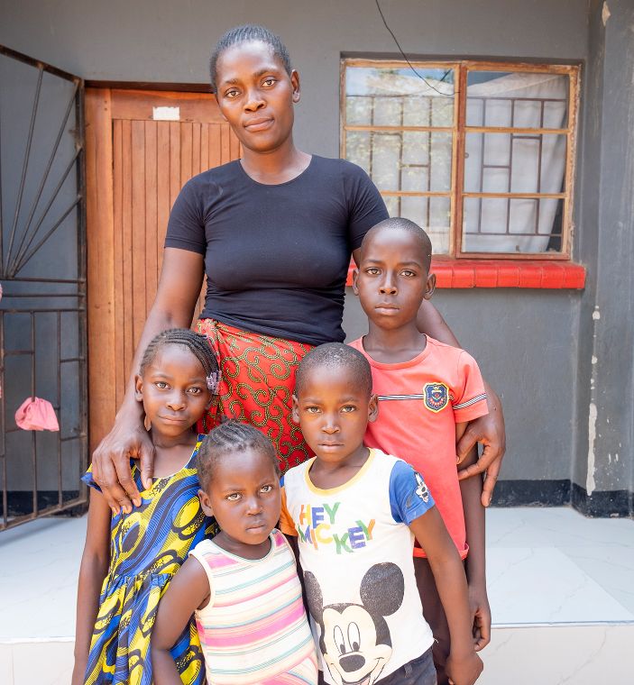 Eine Frau, zwei Mädchen und zwei Jungen stehen vor dem Eingang eines kleinen, grauen Hauses.