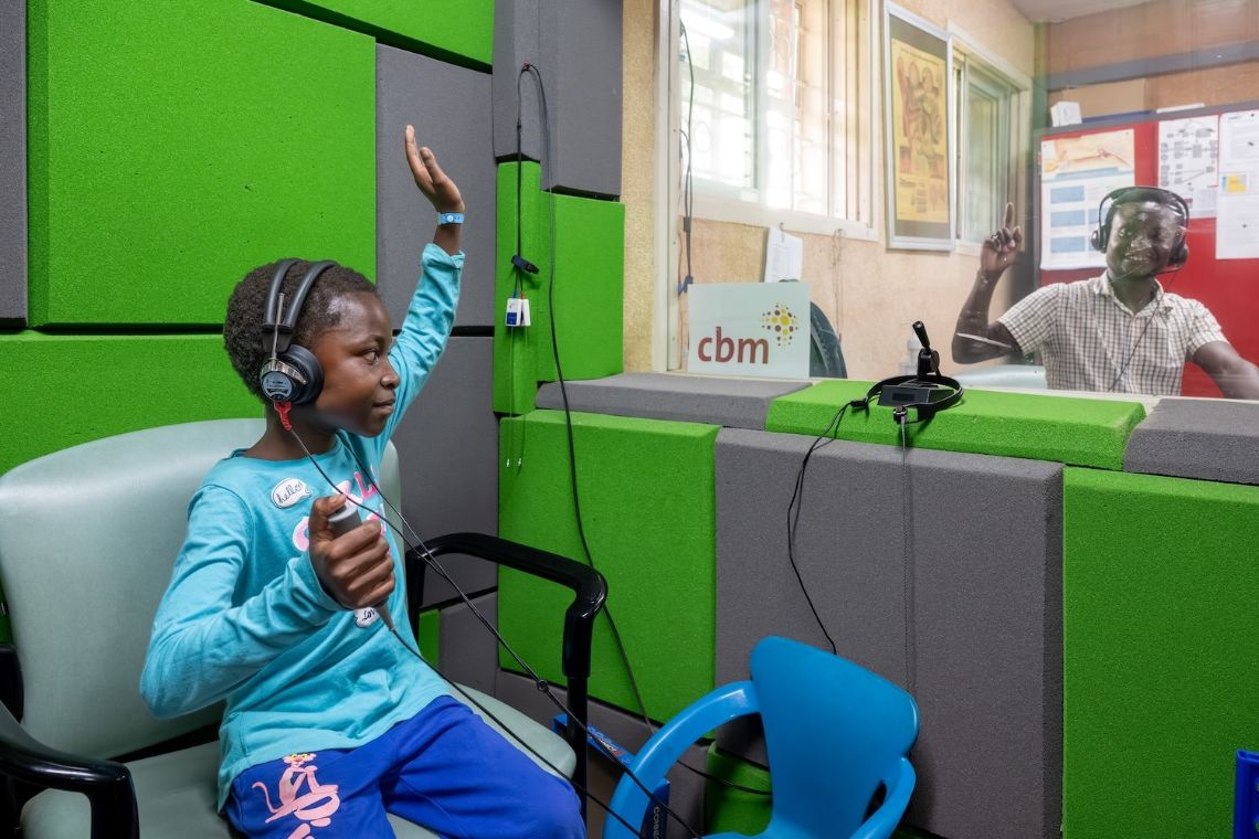 Ein Mädchen mit Kopfhörern hält einen Arm hoch, hinter einer Glasscheibe sitzt ein Mann, der ebenfalls seinen Arm hochhält.