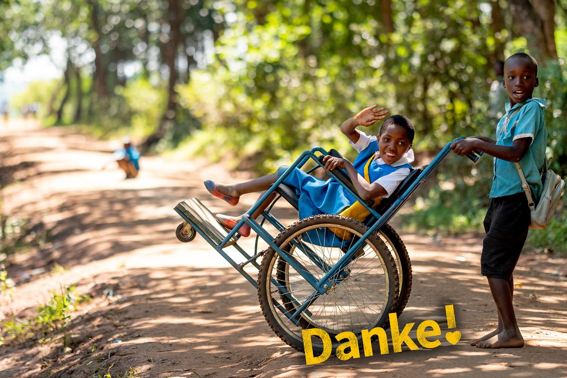 Ein etwa zehnjähriger Junge in Schuluniform schiebt einen Rollstuhl, in dem ein Mädchen sitzt, das ebenfalls eine Schuluniform trägt. Es lacht und winkt.