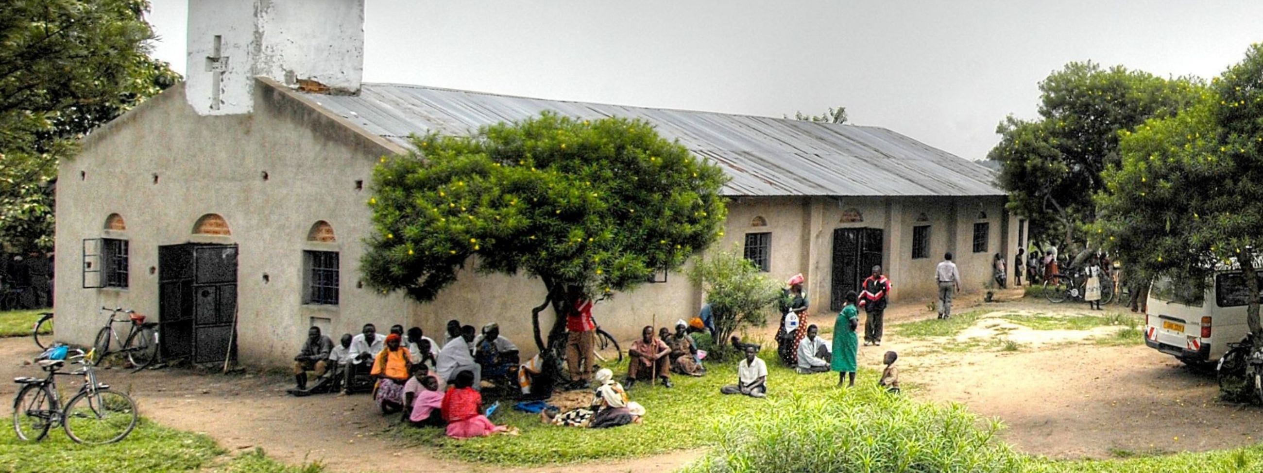 Landschaft mit Kirche, vor der eine Gruppe Menschen in bunter Kleidung sitzt.
