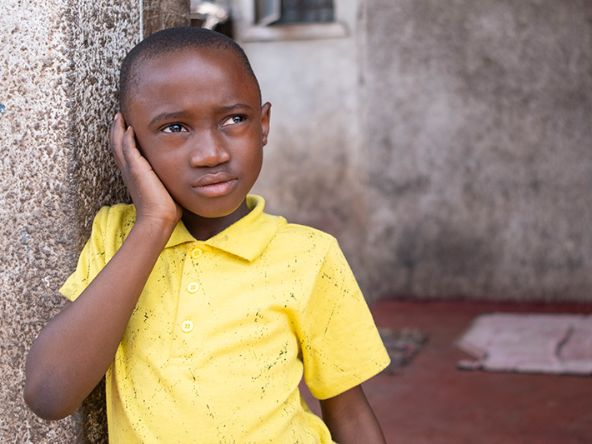Ein etwa achtjähriger Junge lehnt sich an eine Verandamauer; erhält seine rechte Hand an sein rechtes Ohr.