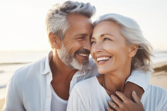Porträtaufnahme eines glücklichen älteren Ehepaares. Der Mann hat seinem Arm um die Frau gelegt, beide lächeln. Im Hintergrund sind Meereswellen und ein Sandstrand zu sehen.