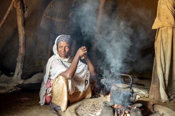Eine Frau mit weiß schimmernden Pupillen sitzt neben einer Feuerstelle.