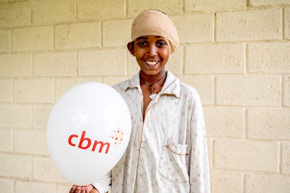 Ein Junge mit Kopfverband hält einen Luftballon in der Hand, auf dem "CBM" steht.