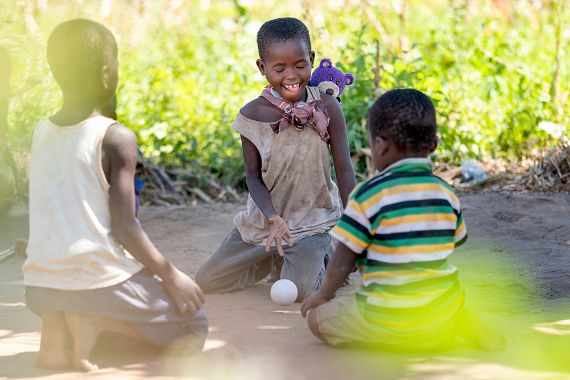 Drei am Boden kniende Kinder spielen mit einem kleinen weißen Ball.