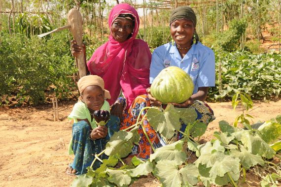 Zwei afrikanische Frauen und ein Kind hocken auf sandigem Boden vor einem Gemüsebeet und halten Gemüse in den Händen.