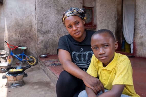 Ein etwa achtjähriger Junge und eine junge Frau sitzen auf einer Veranda vor einer gemauerten Hütte.