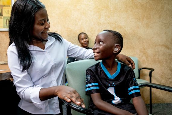 Eine Frau steht neben einem sitzenden Jungen. Im Hintergrund ist eine weitere Frau zu sehen.