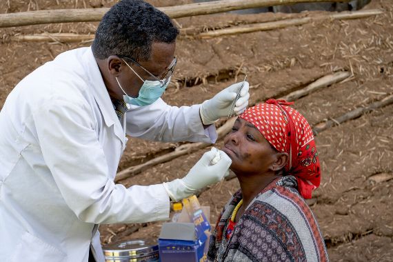 Ein Mann in weißem Kittel führt eine Pinzette zum Auge einer vor ihm sitzenden Frau.