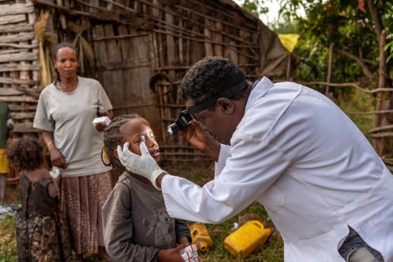 Ein Mann im weißen Kittel leuchtet in die Augen eines kleinen Mädchens. Sie stehen vor einer Hütte.