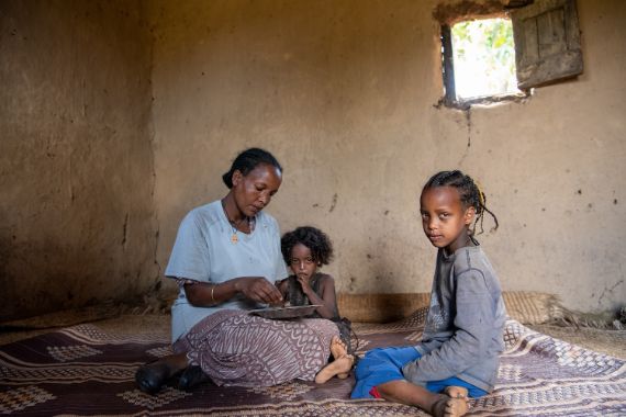 Eine Frau und zwei kleine Mädchen sitzen in einer Lehmhütte auf einem Teppich am Boden. Die Frau hält einen Teller im Schoß und greift mit den Fingern nach etwas, das darauf liegt.