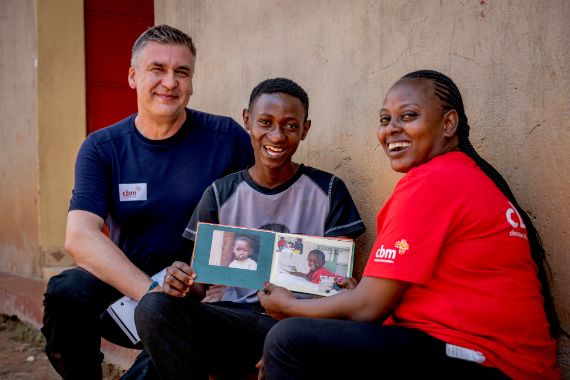 Zwei Männer und eine Frau sitzen auf Vorsprung vor einem Haus. Der Mann in der Mitte ist noch jung. Er hält ein Fotoalbum in der Hand. Auf dem Album ist ein Kinderfoto von ihm. Es zeigt ihn mit einer Lippen-Kiefer-Gaumen-Spalte.