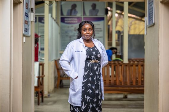 Eine Frau im weißen Kittel über blauweißem Kleid steht in einem Flur, der in eine Halle mit hölzernen Wartebänken mündet.