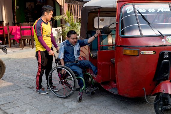 Ein körperbehinderter Mann in Guatemala versucht vom Rollstuhl in ein tuk-tuk einzusteigen.