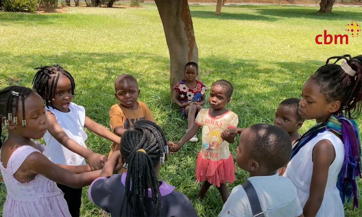 Ein Mädchen sitzt an einen Baum gelehnt. Vor ihr spielen Kinder im Kreis