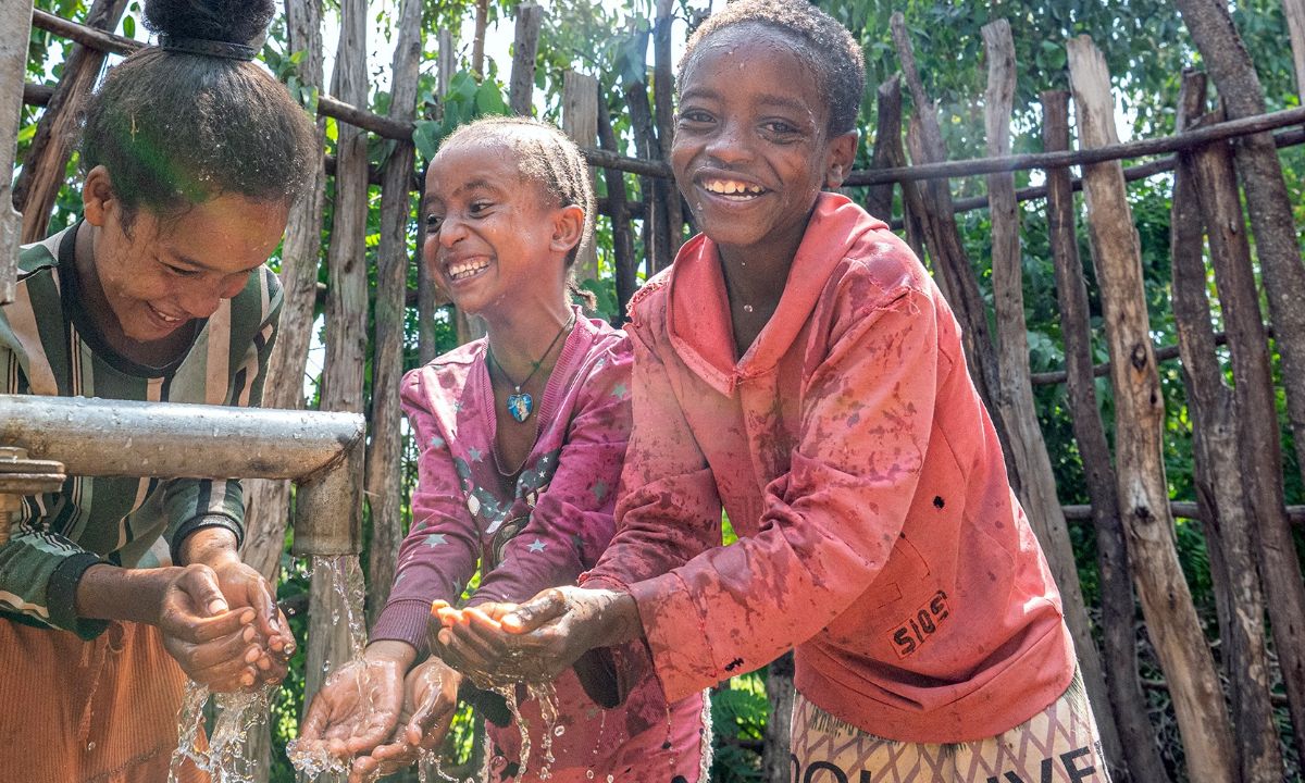 Drei Kinder halten die Hände in den Wasserstrahl, der aus einem Brunnenrohr kommt.