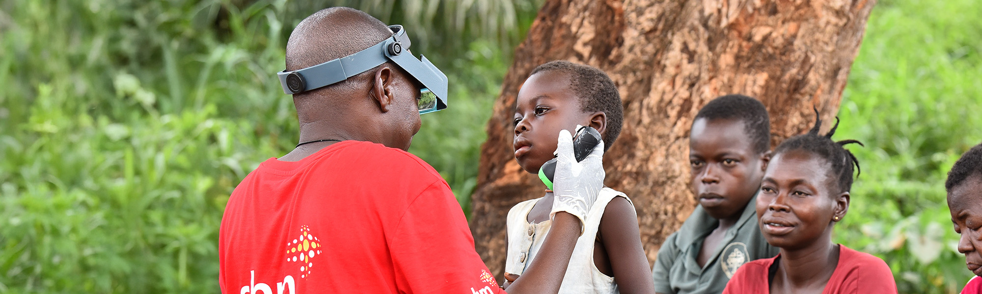 Ein Mann mit Stirnlampe untersucht die Augen eines Kindes, weiter Personen sitzen dabei.